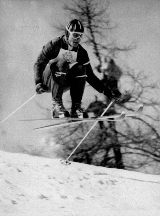 Black and white image of a male skier going over a jump in a downhill race